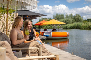 Seijbel-Photography-33-scaled - Stichting Bureau Toerisme - Stichting Bureau Toerisme Two women enjoy colorful cocktails on a wooden dock by a lake under orange umbrellas with a bright blue sky.