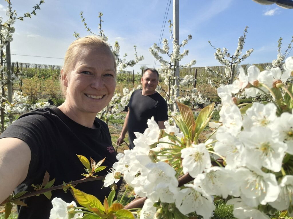 Smiling woman taking a selfie in a blossoming orchard with white flowers on the trees and a man in the background.
