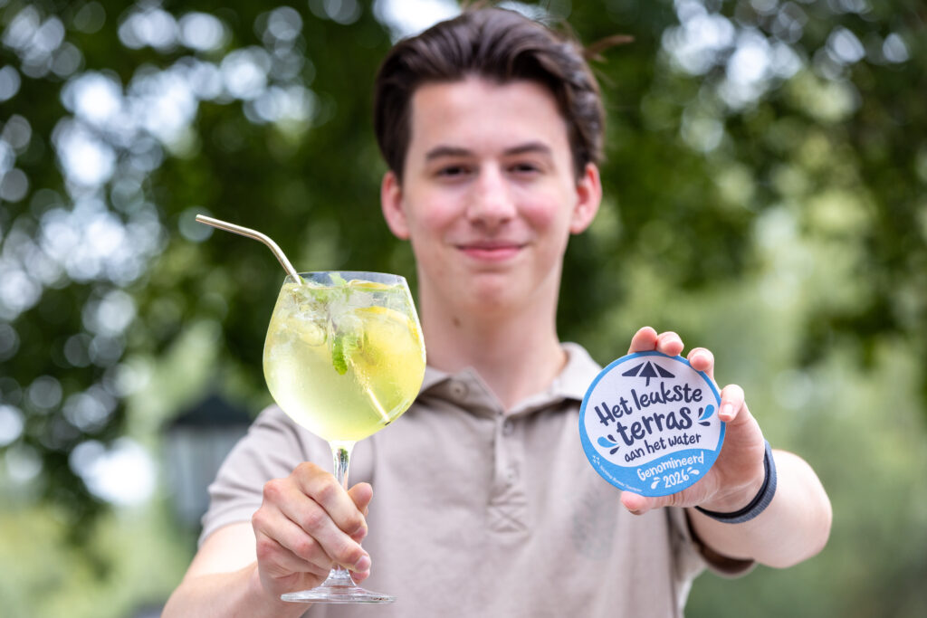 Man smiling and presenting a lime-mint cocktail in a stemmed glass, with a blue circular sign reading 'Het leukste terras aan het water' nearby.