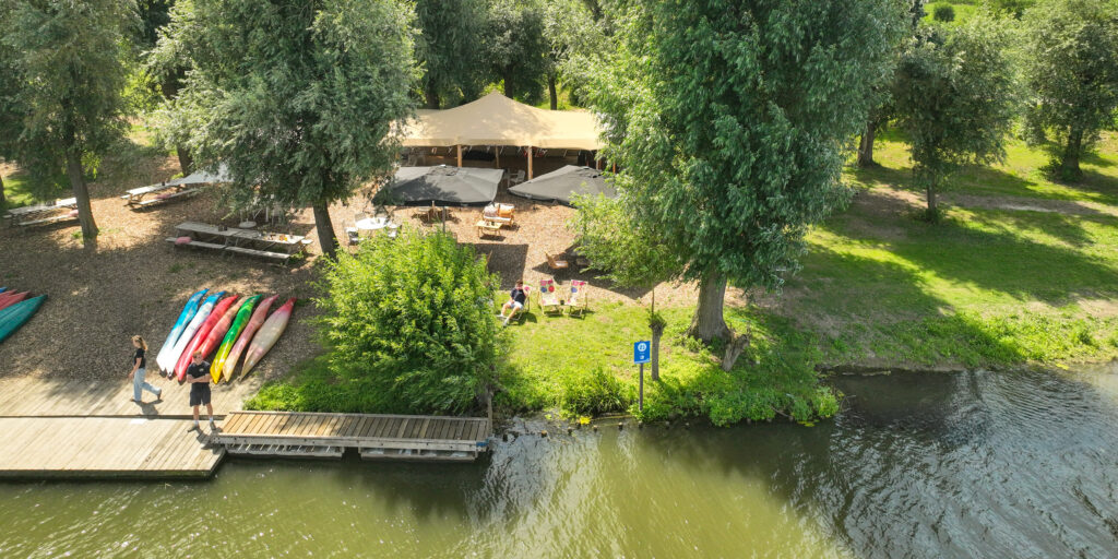 Riverside scene with colorful kayaks lined up on a wooden dock next to a grassy area with a large tent and seating under umbrellas and trees.