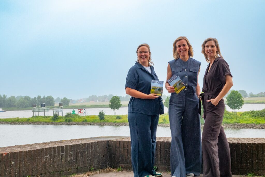 Three women stand by a lake, smiling at the camera and holding brochures.
