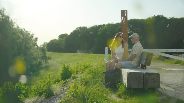 Couple sitting and chatting on a wooden park bench beside a grassy path on a sunny day, with trees in the background.