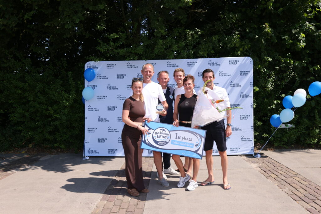 Six people pose outdoors with a first‑place banner and bouquet, in front of a logo backdrop and blue balloons.