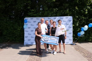 image0 - Stichting Bureau Toerisme - Stichting Bureau Toerisme Six people pose outdoors with a first‑place banner and bouquet, in front of a logo backdrop and blue balloons.